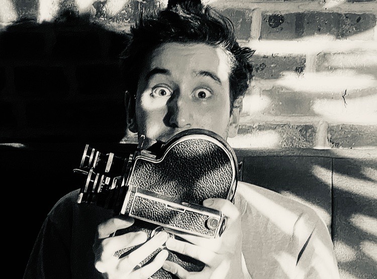 Vasco Sousa Neumann, wide-eyed, holding a 16mm Bolex camera up to his face against a sunlit brick wall. Black and white.